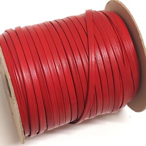 Könnte beinhalten: Eine Spule mit flachem, rotem Lederband, das fest um einen runden Pappkern gewickelt ist. Der Lederstreifen hat eine glatte, gleichmäßige Textur und eine leuchtend rote Farbe, die in mehreren parallelen Schichten über die Spule gewickelt ist.