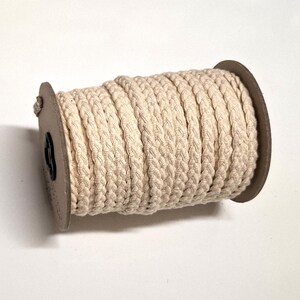 May include: A beige braided cotton rope wound on a cardboard spool. The rope's texture is clearly visible, with individual strands woven together. The spool is set against a white background. Ideal for crafts and macrame projects.