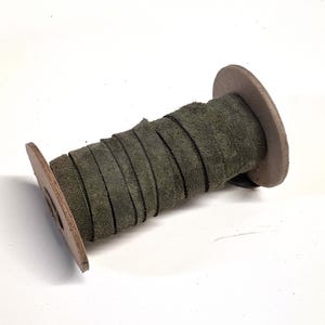 May include: A spool of olive green leather strips, tightly wound around a brown cardboard core. The leather has a textured, suede-like appearance. The spool's edges are visible, showing the layered construction.