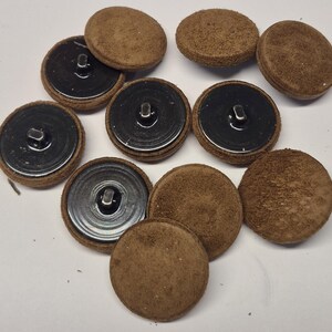 May include: A collection of round buttons featuring a textured brown suede-like top surface and a smooth black metal base with a central shank attachment. The buttons are arranged in a scattered group, showing both the brown fabric sides and the black metal backs.