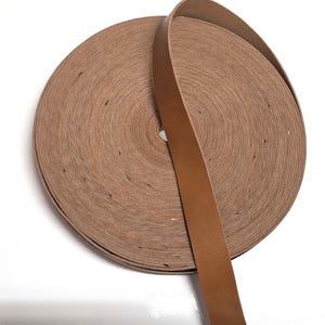 May include: A roll of brown leather strap, approximately 1 inch wide, coiled on a white surface.