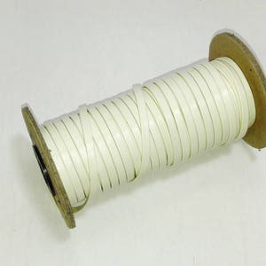 May include: A spool of white leather cord wrapped around a cardboard core. The cord is approximately 6 mm wide.