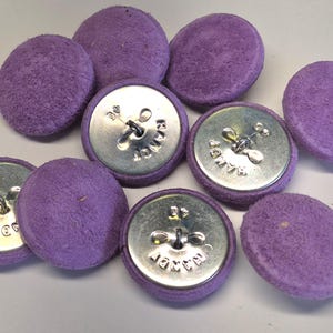 May include: A collection of round buttons covered in a textured, light purple suede-like fabric. The reverse side of the buttons features a metallic silver finish with a central wire shank and the word HANDY stamped into the metal surface.