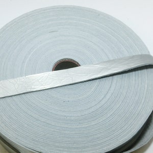 Może przedstawiać: Rolka jasnoszarego sznurka skórzanego, o szerokości około 1,2 cm, z fakturowaną powierzchnią. Sznurek jest nawinięty na szpulę z otworem w środku.