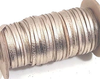 May include: A spool of metallic silver leather cord wrapped tightly around a cylindrical cardboard core. The flat, narrow strip of leather features a reflective, textured surface that catches the light, creating a consistent metallic sheen throughout.