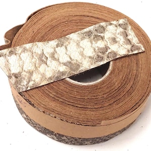 May include: A roll of brown leather trim featuring a textured, metallic silver and grey snakeskin pattern overlay. The material is coiled into a circular shape, displaying the layered edges of the leather strip and the detailed reptilian scale texture.