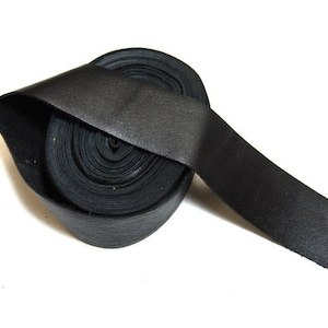 Puede incluir: Una tira de cuero negro, de aproximadamente 2,5 cm de ancho, está enrollada y anudada. La tira está hecha de cuero suave y flexible, y tiene una textura lisa y uniforme.