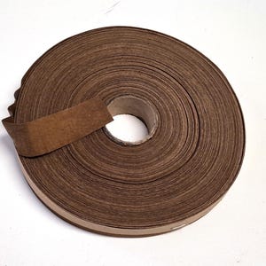 May include: A roll of brown paper tape, unwound slightly to reveal the adhesive side. The tape is tightly wound, showing the edges of each layer. The roll sits on a white surface.