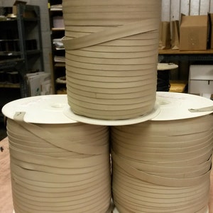 Puede incluir: Tres carretes de cinta de tela beige enrollados en carretes de cartón blanco. La cinta es un material plano y tejido con una textura sutil.