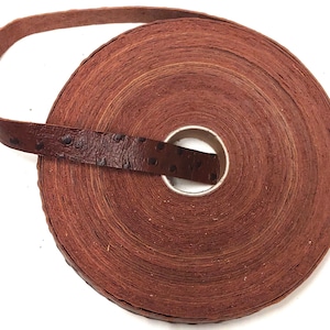 May include: A continuous roll of dark brown leather strapping featuring a distinct textured surface with small, evenly spaced indentations. The leather strip is wound tightly around a central silver-coloured metal core, with one end extending outwards.