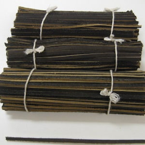 Puede incluir: Una pila de tiras de cuero negro y marrón atadas con una cuerda blanca. Las tiras tienen aproximadamente 1,2 cm de ancho y están apiladas a unos 7,5 cm de alto.