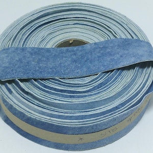 May include: A coiled roll of light blue suede leather strip with a fuzzy texture. The strip is wound into a tight circular shape and secured with a tan paper band that has handwritten text on the side, showing a consistent blue colour throughout.