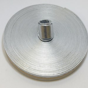 Puede incluir: Un flexómetro metálico plateado enrollado, con un centro cilíndrico de metal. El flexómetro es circular, con anillos concéntricos. El centro es un casquillo hexagonal plateado. Sobre un fondo blanco.