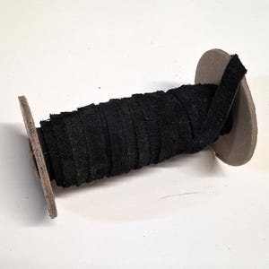 May include: A spool of black fabric trim. The trim is tightly wound around a cardboard and wood spool. The fabric appears to be a textured material, possibly suede or a similar fabric. The spool is set against a white background.