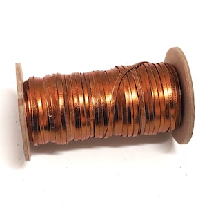 May include: A spool of copper-coloured leather cord wrapped around a cardboard spool. The cord is thin and has a shiny finish.