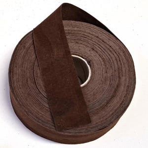 Può includere: Un rotolo di nastro marrone scuro, ben avvolto, con un pezzo di nastro che si estende verso l'esterno. Il nastro sembra essere fatto di un materiale morbido, simile al tessuto. Il rotolo è centrato su uno sfondo bianco.