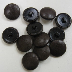 Peut inclure: Un groupe de boutons en cuir marron avec des centres en plastique noir. Les boutons sont éparpillés sur une surface blanche.