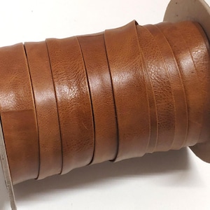 May include: A spool of flat, tan-colored leather cord wrapped tightly around a circular cardboard core. The leather strip has a smooth, consistent texture and a warm brown tone, wound in multiple overlapping layers across the cylindrical spool.