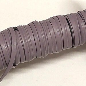 May include: A spool of flat, mauve-colored leather cord wrapped tightly around a cardboard cylinder. The cord has a smooth, matte finish and is wound in multiple layers, showing a consistent width throughout the entire length of the spool.
