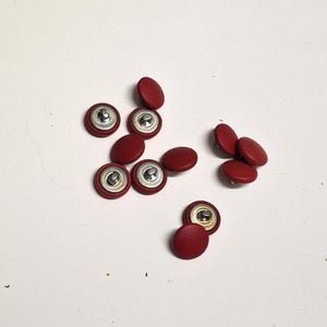 Peut inclure: Plusieurs boutons ronds recouverts de cuir rouge avec des dos en métal argenté. Les boutons sont légèrement bombés et sont dispersés sur une surface blanche.