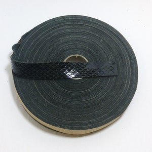 Może przedstawiać: Rolka czarno-zielonej taśmy tekstylnej z wzorem skóry węża. Taśma jest zwinięta i ma beżowy brzeg.