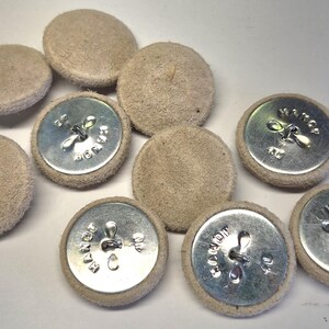 May include: A collection of round buttons featuring a beige suede-like fabric covering on the front and a silver-toned metal back. The metal backs are stamped with the text "HANDY" and "MADE" along with a central metal loop for attachment.