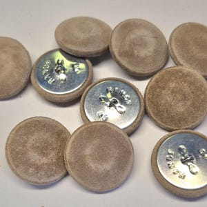 May include: A collection of round buttons featuring a soft, light brown suede-like fabric covering on the front. The reverse side of the buttons is made of polished silver-colored metal with a central shank and the text RAMON 48 embossed on it.