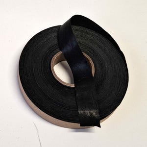 May include: A roll of black satin ribbon. The ribbon is tightly wound around a cardboard core, with a length of ribbon extending from the roll. The ribbon has a smooth, shiny texture and is a deep, solid black color.