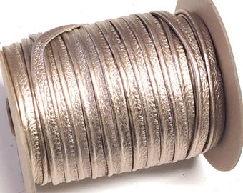 Puede incluir: Una bobina de cordón de cuero color champán metalizado enrollado firmemente alrededor de un núcleo de cartón. El cordón tiene una textura plana y lisa con un brillo sutil, mostrando múltiples capas enrolladas de forma uniforme.