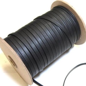 May include: A spool of flat black cord wrapped around a cylindrical cardboard core. The cord is wound tightly in a continuous spiral pattern, creating a uniform texture across the surface of the spool. A single loose strand extends from the base.
