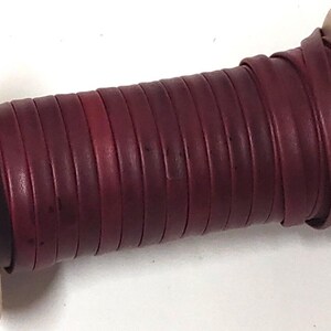 May include: A spool of deep burgundy leather cord wrapped neatly around a light tan cardboard cylinder. The flat, smooth leather strip is wound in tight, uniform layers, showing a consistent dark red colour throughout the entire length of the cord.