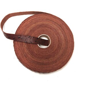 Może przedstawiać: Rolka brązowej wstążki skórzanej z fakturowanym wzorem przypominającym skórę strusia. Wstążka jest zwinięta i związana pętlą.