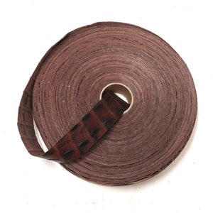 Może przedstawiać: Rolka brązowej wstążki skórzanej z wzorem krokodyla. Wstążka jest zwinięta i związana kawałkiem tej samej skóry.