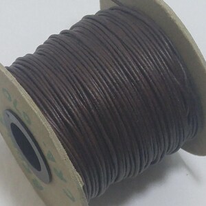 May include: A spool of dark brown leather cord wrapped tightly around a circular cardboard core. The cord has a smooth, rounded texture and is wound in multiple layers across the width of the spool, which features a central black hole.