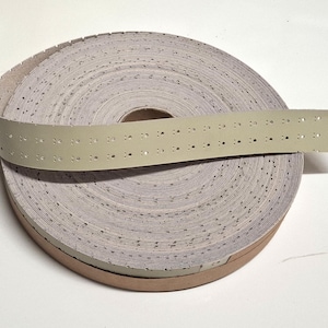 May include: A roll of perforated paper tape, possibly for a vintage machine. The tape is a light grey colour with small, evenly spaced holes. A section of the tape extends from the roll, showing the perforations.