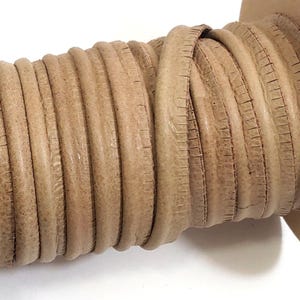 May include: A spool of light brown, round leather cord wrapped tightly around a cardboard core. The cord features a visible, textured grain and a consistent cylindrical shape, wound in multiple neat layers across the length of the spool.