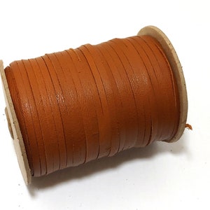 May include: A spool of brown leather cord wrapped around a cardboard tube. The cord is thin and flat, and it is perfect for crafting projects.