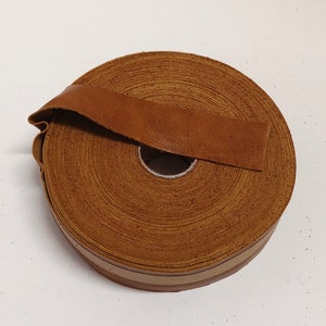 Puede incluir: Un rollo de cordón de cuero marrón con un borde beige. El cordón está enrollado y atado con un trozo del mismo cuero.