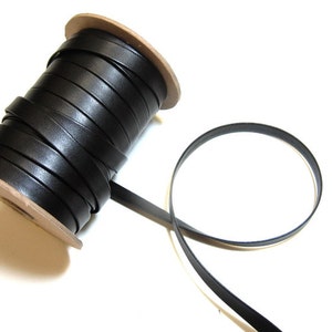 May include: A spool of black leather cord, wound around a cardboard core. The cord is about 1/4 inch wide and is shown unfurling from the spool.