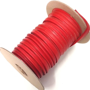 May include: A spool of bright red zip tape featuring a continuous coil design wrapped around a light brown cardboard core. The zip tape has a smooth, flat surface with visible teeth along the edges, wound tightly in multiple layers.