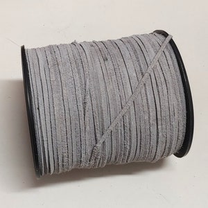 May include: A spool of gray suede cord, wound tightly on a black plastic bobbin. The cord is made of thin strips of leather, woven together to create a durable and flexible material.
