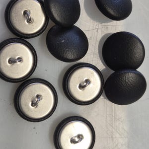 Op de afbeelding: Een verzameling zwarte, ovaalvormige knopen met een getextureerd oppervlak. Elke knoop heeft een zilverkleurige metalen achterkant met een lus om te naaien. De knopen liggen op een lichtgekleurd oppervlak.