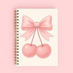 Puede incluir: Un cuaderno con espiral con una cubierta rosa. La cubierta presenta una ilustración de acuarela de un lazo rosa y dos cerezas rosas. El cuaderno tiene una encuadernación en espiral dorada.