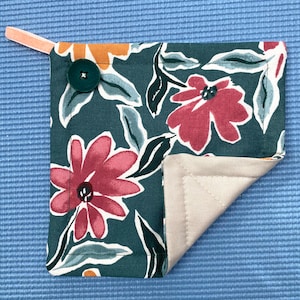 May include: A square, floral-patterned pot holder with a dark teal background and large pink flowers. The pot holder has a beige interior, a green button, and a peach-colored loop for hanging.