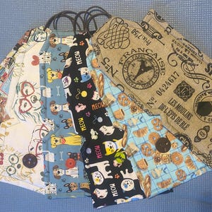 May include: A collection of fabric tote bags with various patterns. Designs include dogs, cats, maps, and Parisian-themed prints. Each bag has a black handle and a button closure. The bags are arranged on a blue surface.