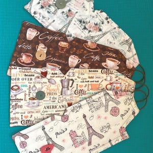 May include: A collection of handmade fabric face masks with various patterns. Designs include coffee-themed prints with coffee cups and text, and a Parisian theme with Eiffel Towers and floral accents. Each mask has elastic ear loops.