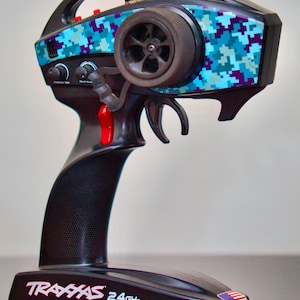 Könnte beinhalten: Eine schwarze Fernbedienung mit einem blauen und violetten Tarnmuster. Die Fernbedienung hat einen roten Abzug und eine kleine amerikanische Flagge auf der Basis. Der Text "Traxxas 2.4GHz" ist auf der Basis aufgedruckt.