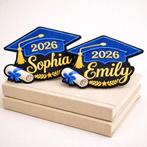 Può includere: Due toppe di laurea con cappelli blu, l'anno 2026 e i nomi "Sophia" ed "Emily" in oro. Ogni toppa include un diploma arrotolato legato con un nastro blu. Le toppe sono poste su una pila di due libri beige.