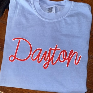 May include: A light blue t-shirt with the name "Dayton" in red script. The shirt is folded and appears to be made of a soft material. The tag reads "Comfort Colors".