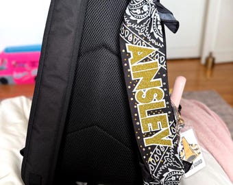 May include: A black backpack with a padded mesh back panel and adjustable straps. One strap features a black and white paisley fabric wrap with the name AINSLEY in gold glitter lettering, accented with small gold studs and a cheer megaphone tag.